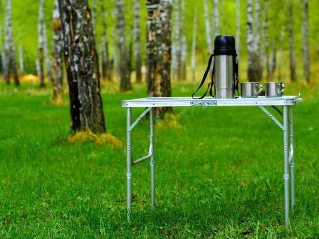 camping table with thermos and two cups on top sitting in the woods.