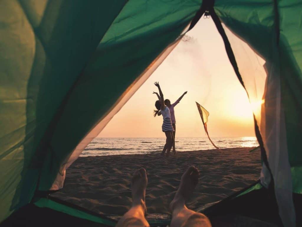 person in a green tent on a sandy beach while doing some pismo beach camping looking out to see two people flying a kite near the water.