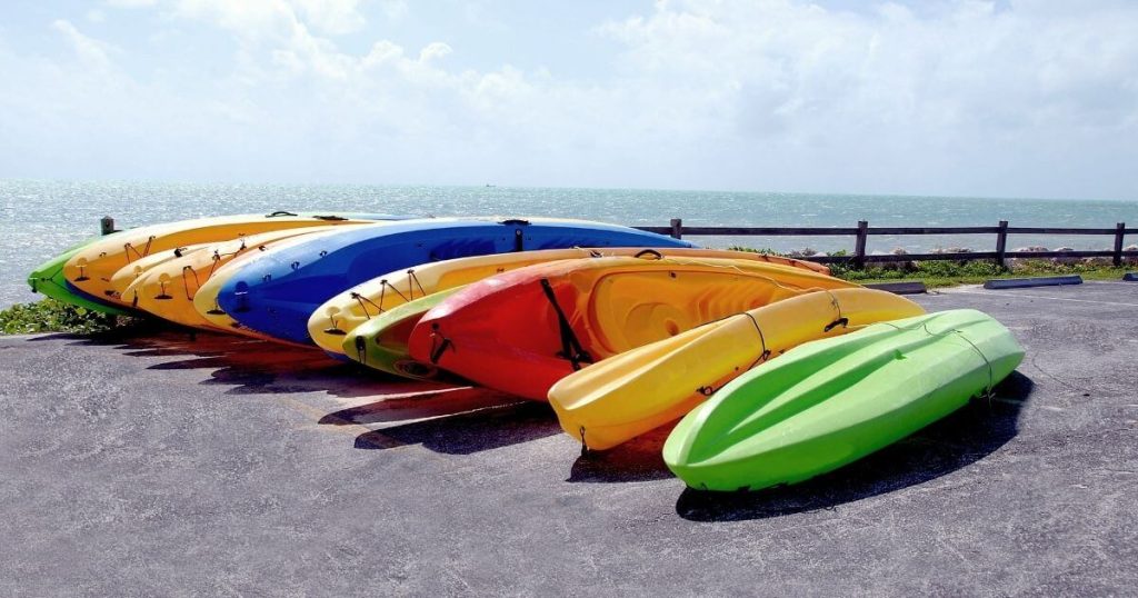 A bunch of the best kayaks for camping lined up on the ground near a body of water.